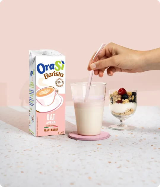 Confezione di OraSì Barista Avena accanto a una tazza di cappuccino e una ciotola di yogurt con frutti di bosco su sfondo rosa