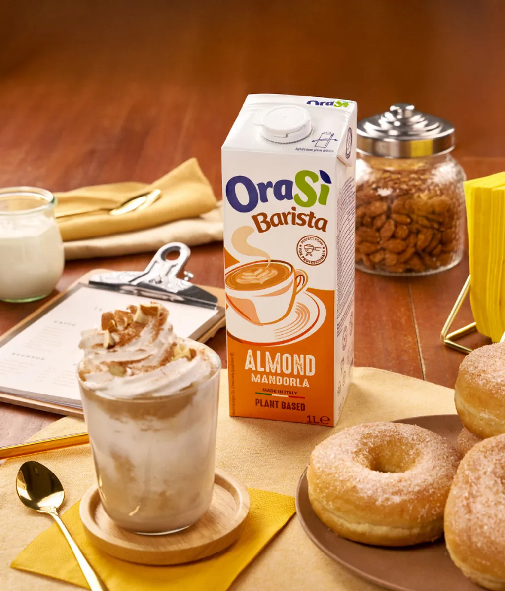 OraSì Barista bevanda alla mandorla su un tavolo su una tovaglietta beige con un bicchiere con panna, con un piatto con dei bomboloni e un cucchiaino per colazione