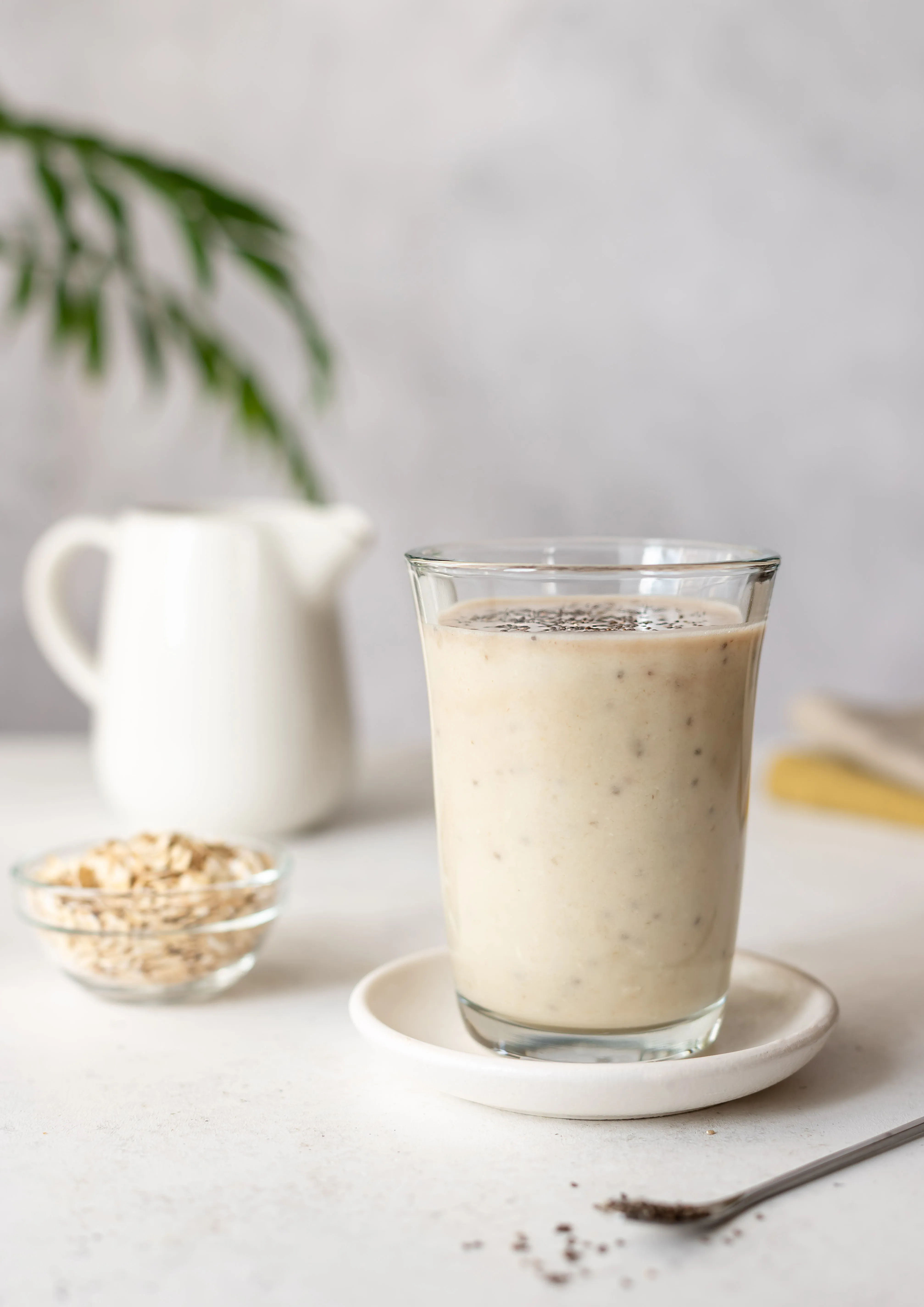 A glass of oat smoothie next to a small bowl of rolled oats and a white pitcher.