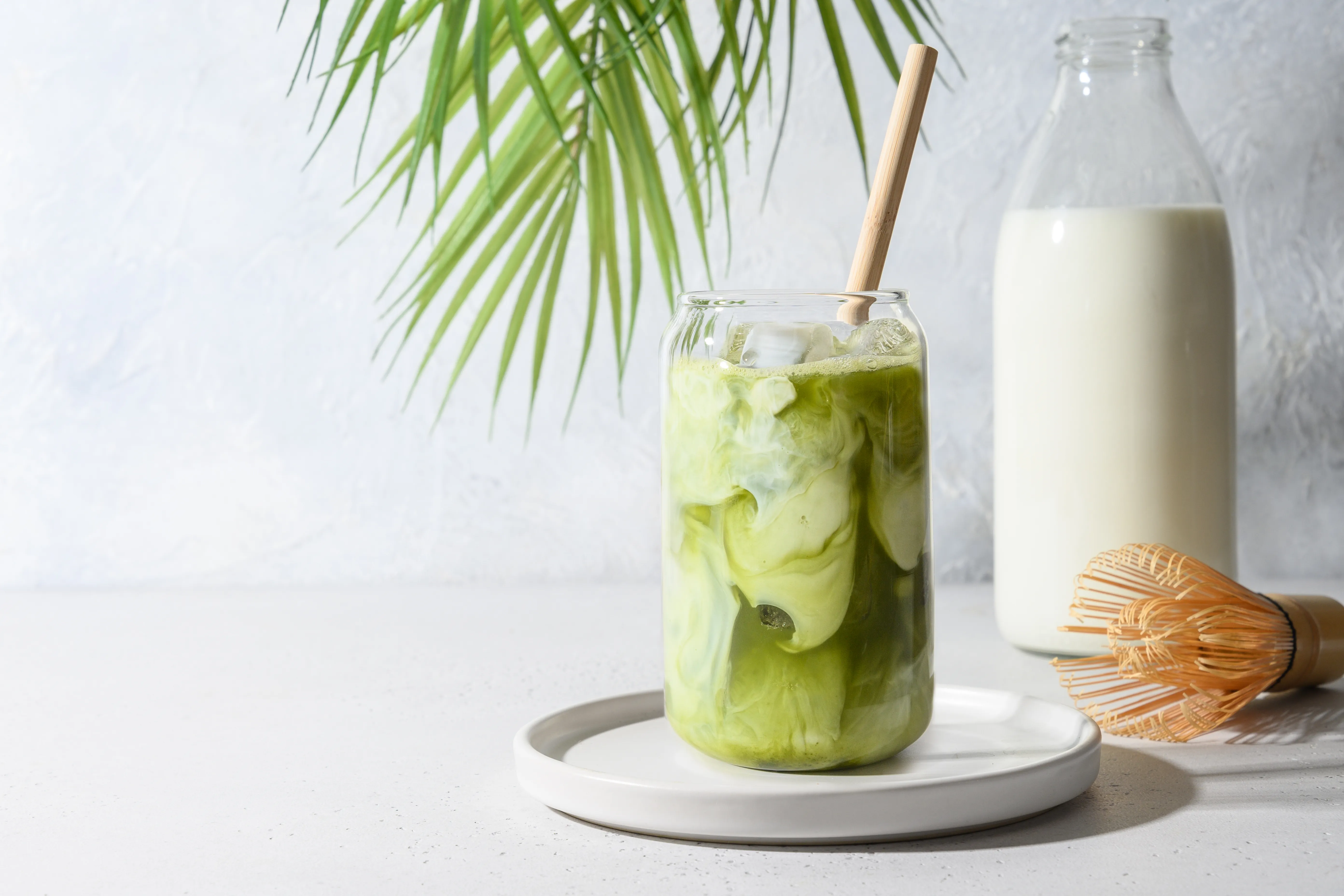 A glass of iced matcha latte with ice cubes and a straw next to a milk bottle.