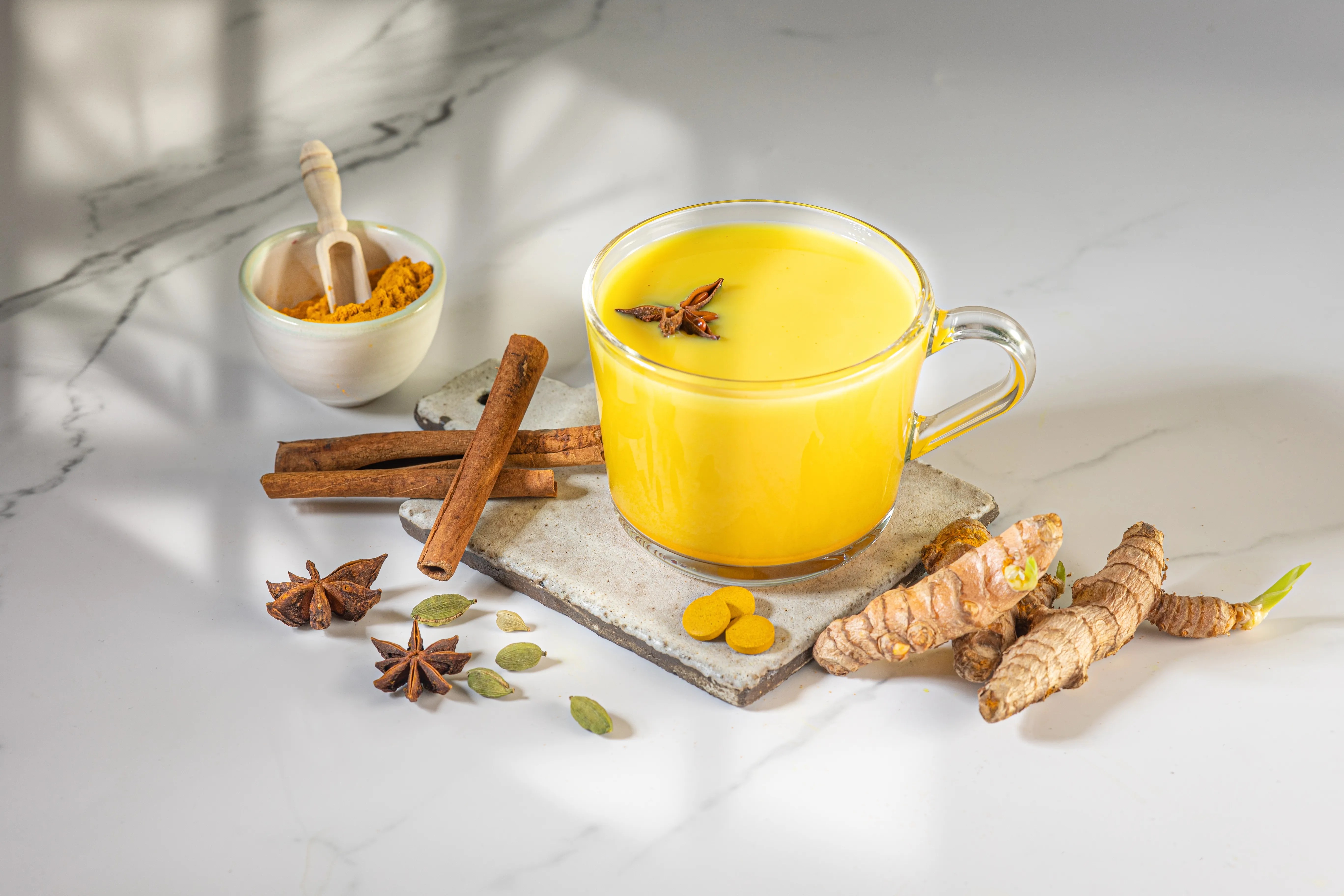 Glass mug of turmeric Golden Milk surrounded by cinnamon sticks, star anise, and fresh turmeric root.
