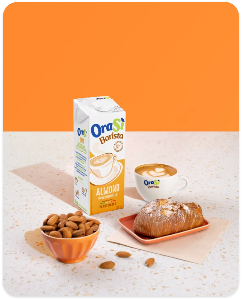 OraSì Barista bevanda alla mandorla con mandorle in una ciotola e un croissant su un tavolo su sfondo arancione